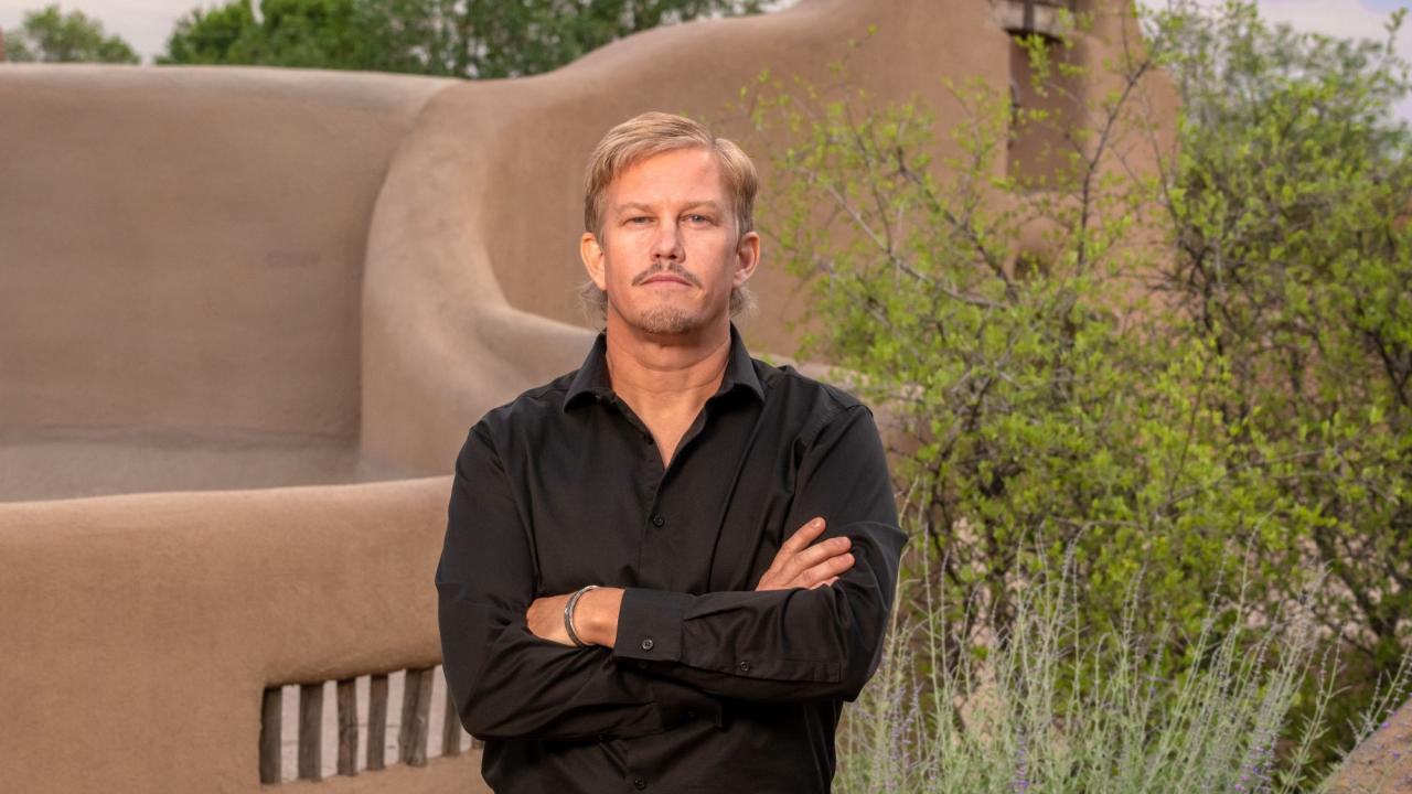 A man stands near an adobe structure with crossed arms